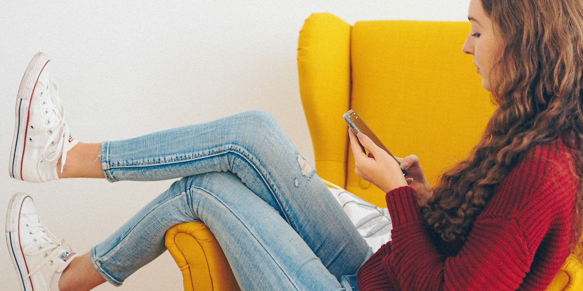 Teenager scrolling on phone Teenage girl curled up in a yellow armchair scrolling on her phone