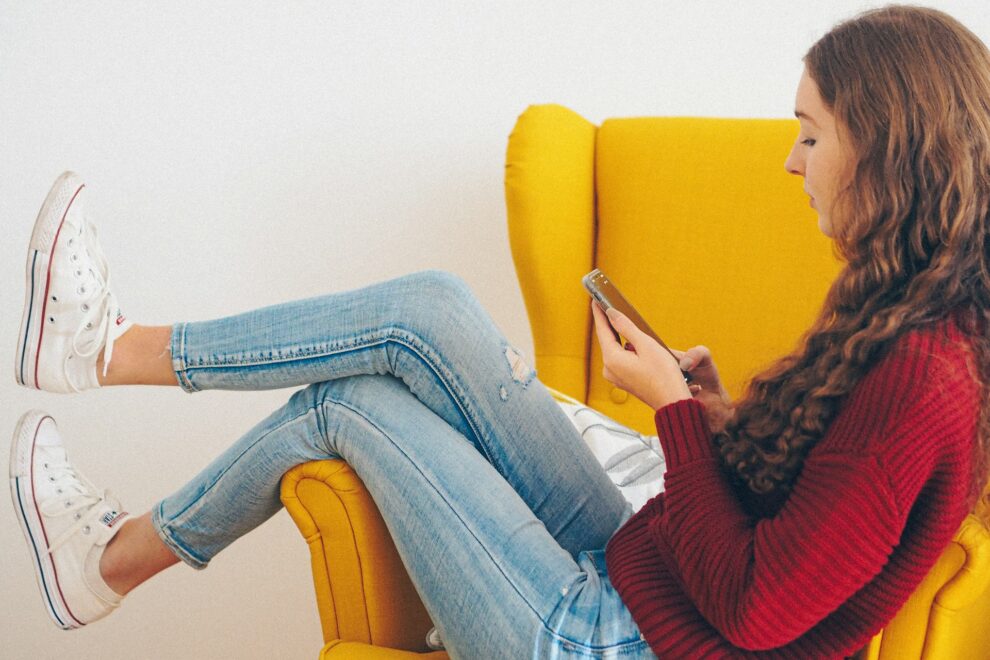 Teenage girl curled up in a yellow armchair scrolling on her phone